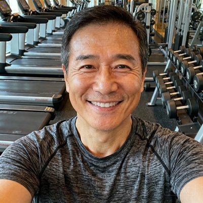 张伟的正面半身照，一位35岁左右穿着运动T恤的男子在健身房自拍，背景是跑步机和哑铃架，面带自信微笑